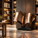 Abstract metal open book sculpture on a black base displayed in an indoor hallway with bookshelves and polished marble floors, adding an elegant artistic touch to luxury library and interior spaces.