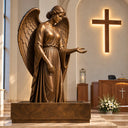 Bronze angel sculpture with feathered wings and an open hand displayed beside a wooden cross inside a contemporary church.