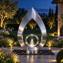 Illuminated by landscape lighting at dusk, this brushed stainless steel teardrop fountain sculpture frames an inner ring with rising water, offering a composed focal point for a villa garden.