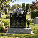 Dedicated to a beloved memory, this custom grave monument with a detailed elephant carving rests among headstones in a lush cemetery background.
