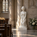 Classical white marble praying woman statue with a draped robe and flowers, displayed inside a church or memorial hall with wooden pews and stained glass windows, suitable for indoor religious and memorial decor.

