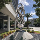 Contemporary abstract metal sculpture with flowing reflective stainless steel forms displayed in a landscaped luxury villa courtyard.