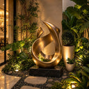 Framed by planting, the gold-finished bronze sculpture pairs crossing ribbons with a dark rectangular base within a villa courtyard.