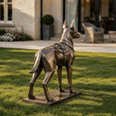 A custom bronze working dog sculpture featuring a faceted geometric design, set on a gravel patch with a stone building in the distance.