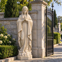 Standing beside an iron gate, this Egyptian Beige marble Virgin Mary statue features a prayerful pose, serving as a Catholic garden sculpture.