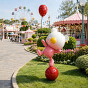 A fiberglass bear sculpture with a white head, pink body and red balloon balances on a red sphere in an amusement-park setting.