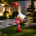Distinguished by its lifted leg and glossy pink-and-white body, this fiberglass bear sculpture carries a red balloon within a landscaped courtyard setting.