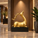 Gold reclining gazelle sculpture with long horns on a black base, placed in a luxury lobby with marble floors and wood paneling, ideal for high-end interior art decoration and hotel design.