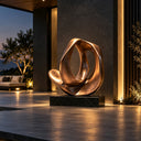 Golden abstract loop sculpture on a black pedestal displayed in a modern indoor lobby or entrance at dusk with warm lighting, adding a contemporary artistic touch to luxury interior decor.
