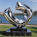 Large abstract stainless steel sculpture with flowing mirror-polished curves displayed on a black marble pedestal beside the waterfront.