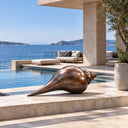 A monumental bronze conch shell sculpture with a smooth spiraled form, sitting on stone steps for a coastal garden setting.
