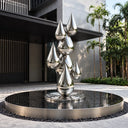 Large mirror-polished stainless steel water drop sculpture displayed on a circular platform outside a modern architectural entrance