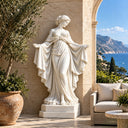 Classical white marble goddess statue with a flowing robe and outstretched arms on a seaside terrace with olive trees, sea view, and outdoor furniture, suitable for luxury villa patio and garden decoration.