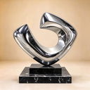 Abstract stainless steel sculpture with an asymmetrical open-loop design, polished silver finish, and black marble pedestal against a beige background.
 Copy