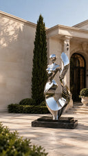 A mirror polished stainless steel abstract female sculpture reflecting the surrounding garden, placed on a black marble base.