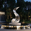 A mirror polished stainless steel sculpture exhibiting an upward spiral form, placed on a circular base with surrounding water reflections in a dark outdoor setting.