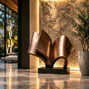 Modern abstract bronze sculpture shaped like an open book, placed in a luxury lobby with marble floors and a textured stone wall, ideal for high-end interior art decoration.