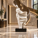 Modern white abstract figure sculpture placed beside a spiral staircase in a luxury hotel lobby, used as artistic interior decor to enhance the elegant and upscale atmosphere of the space.