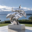 Large polished stainless steel dolphin artwork displayed in a landscaped outdoor architectural setting