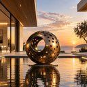 Perforated bronze sphere sculpture BB-013 beside a reflecting pool at sunset