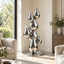 Silver abstract water drop sculpture with stacked teardrop shapes displayed in a modern living room near large windows and a sofa, adding a contemporary artistic touch to luxury interior decor.