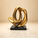 Studio shot of a modern abstract gold sculpture with a twisted organic loop design on a black base against a beige background, showcasing its polished metallic texture for art decor product display and reference.


