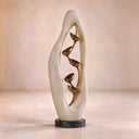 Resting on a dark stone base, this marble and bronze sculpture reveals pale stone veining and warm brown flower forms in a studio setting.