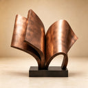Studio shot of a modern bronze open book sculpture with a polished metallic finish on a black base against a beige background, showcasing its elegant curves and texture for art decor product display and reference.
