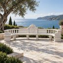 This white marble outdoor bench combines a scalloped backrest with carved stone supports, delivering elegant seating for a seaside terrace.

